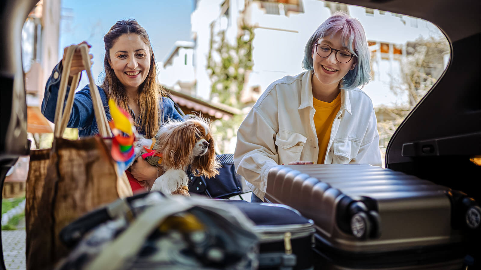 duas raparigas com um cão ao colo a encherem o porta-bagagem do carro para irem de férias